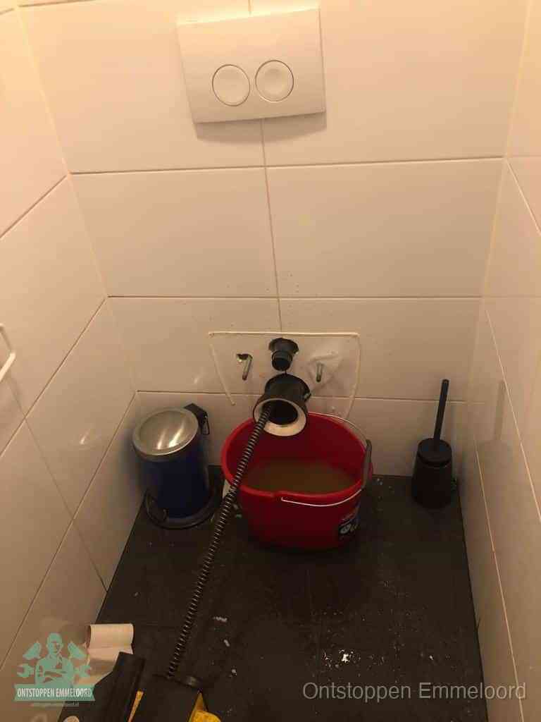 Een ontstopte wand-wc met verwijderde toiletpot waar een ontstoppingsveer in de wanduitgang verdwijnt, het achterhalen waar de verstopping zit in Emmeloord