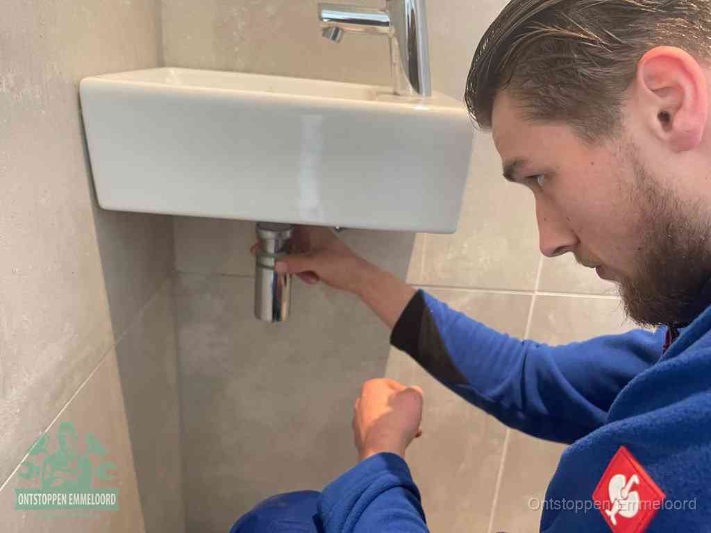 Een monteur draait met de hand de zwanenhalssifon onder een fonteinwasbak in de toiletruimte los om de bron van rioollucht in Emmeloord te vinden
