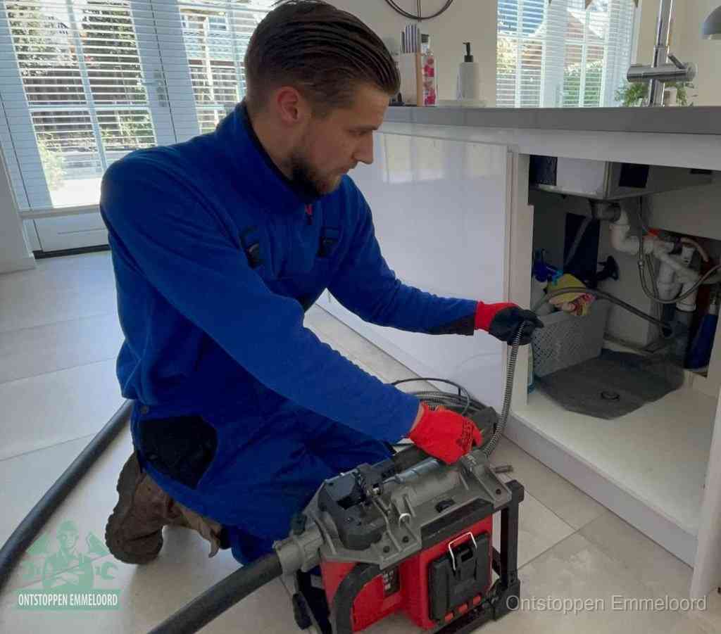 Een monteur met rode handschoenen knielt in een keuken en voert een elektrische ontstoppingsveer in tijdens het professioneel afvoer ontstoppen in Emmeloord